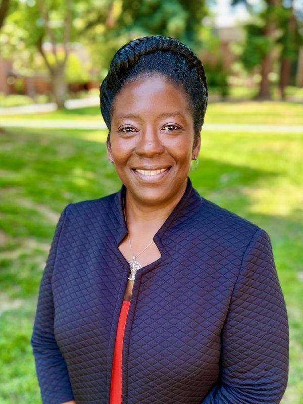 The photo shows the smiling face of Dr. Tanisha Sparks. she is wearing a dark blue jacked over an orange shirt while standing in the shade of a tree on the quad at the Sacramento campus.